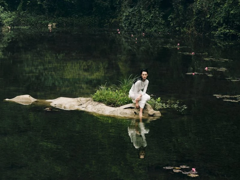 Asian woman in white clothes sits barefoot on rock in a lake with lily pads surrounded by green trees in a natural setting.