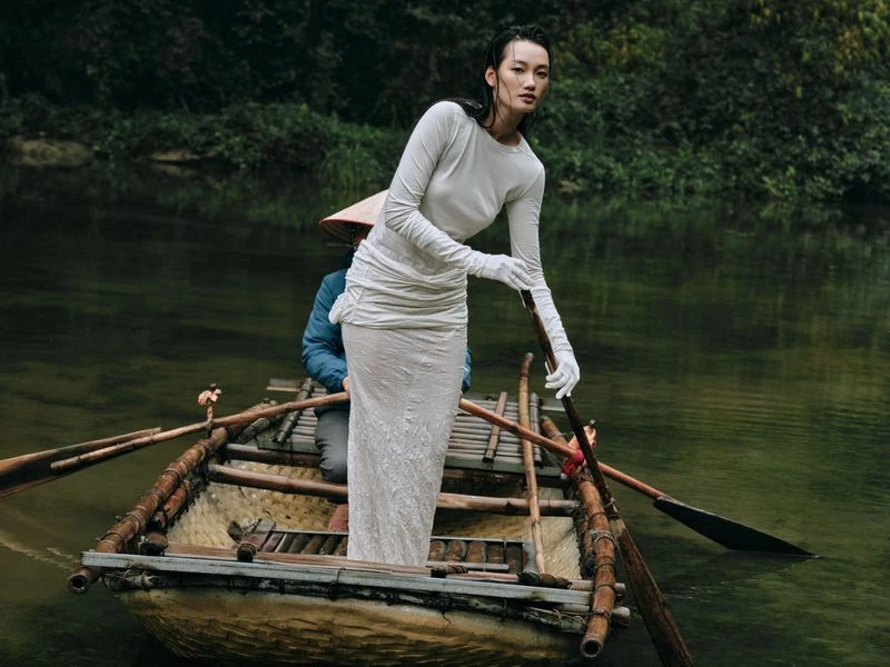 Elegant woman in white dress on a wooden boat in a river. Man in traditional hat rows the boat. Lush green trees line the riverbank.