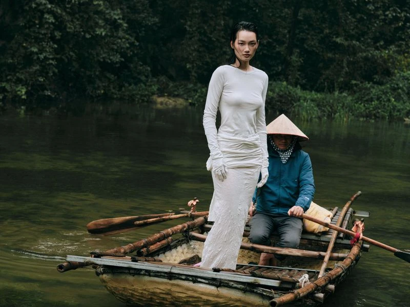 Woman in a long white dress poses on a wooden boat with a man wearing a conical hat, floating on a river surrounded by lush trees.