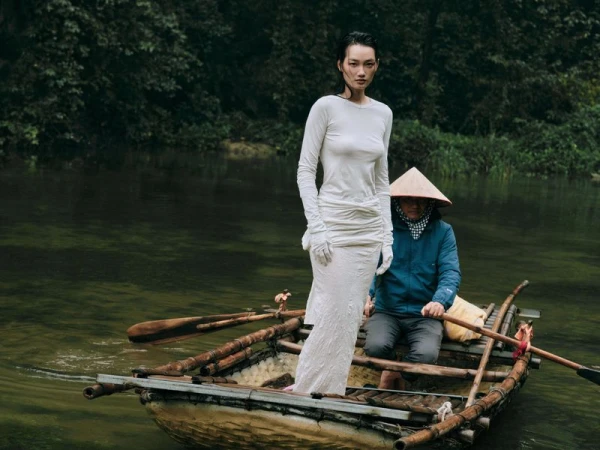 Woman in a long white dress poses on a wooden boat with a man wearing a conical hat, floating on a river surrounded by lush trees.