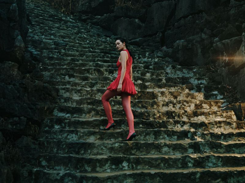 Woman in red dress ascends a stone staircase outdoors surrounded by rocky cliffs, lit with subtle light flare for a fashion editorial.
