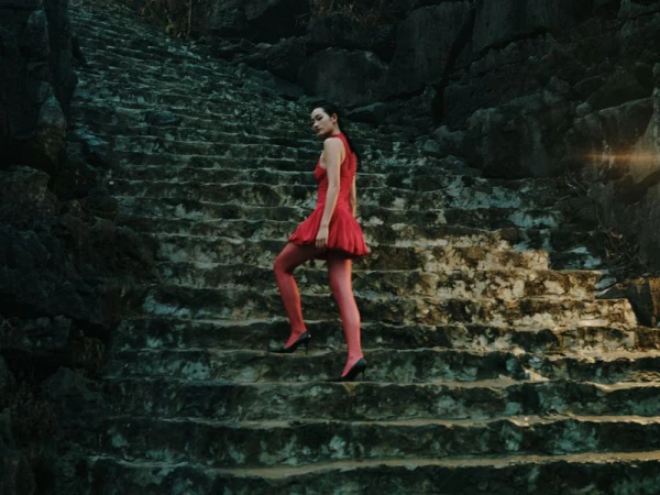Woman in red dress ascends a stone staircase outdoors surrounded by rocky cliffs, lit with subtle light flare for a fashion editorial.