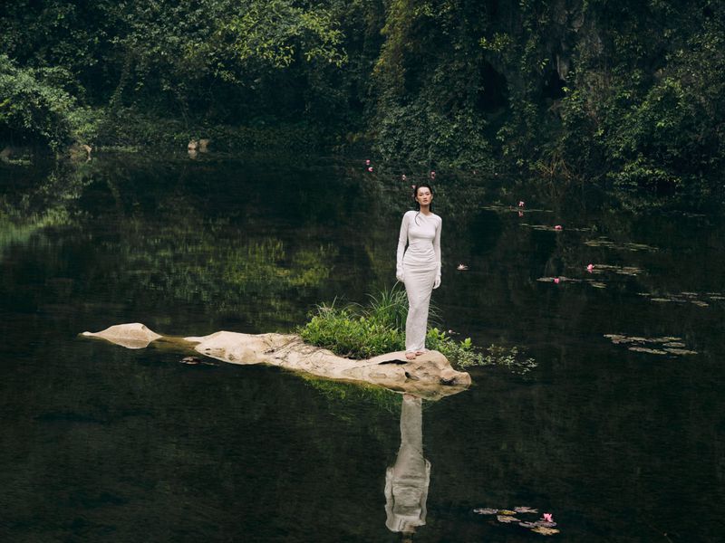 Woman in a long white dress standing on a small rock island in the middle of a lake, surrounded by lush green vegetation and water lilies.