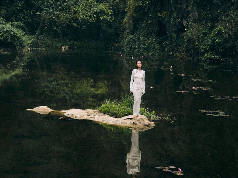 Woman in a long white dress standing on a small rock island in the middle of a lake, surrounded by lush green vegetation and water lilies.