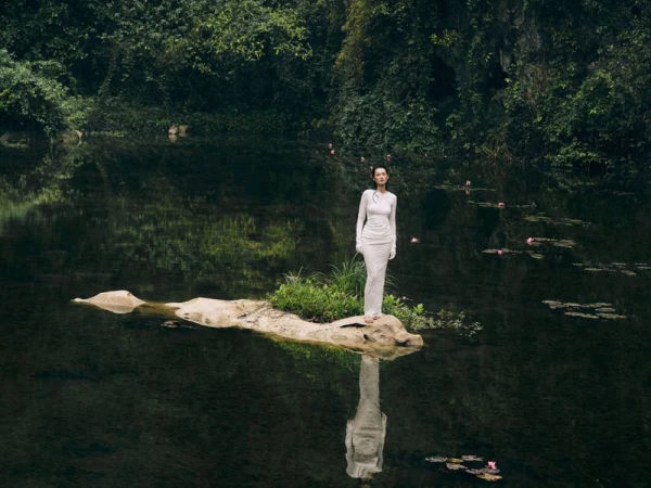Woman in a long white dress standing on a small rock island in the middle of a lake, surrounded by lush green vegetation and water lilies.
