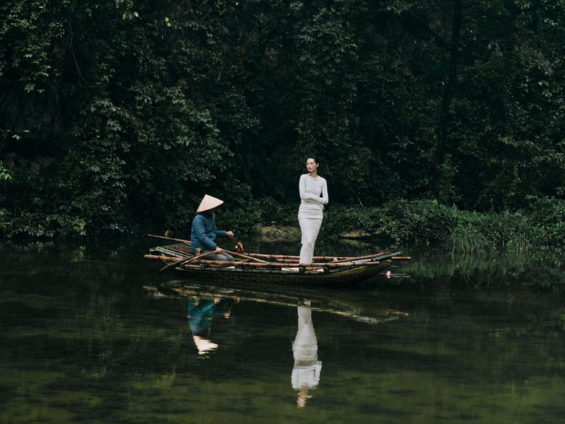 Woman in white dress stands in bamboo boat on water near seated man wearing a conical hat in a lush, green, outdoor setting.