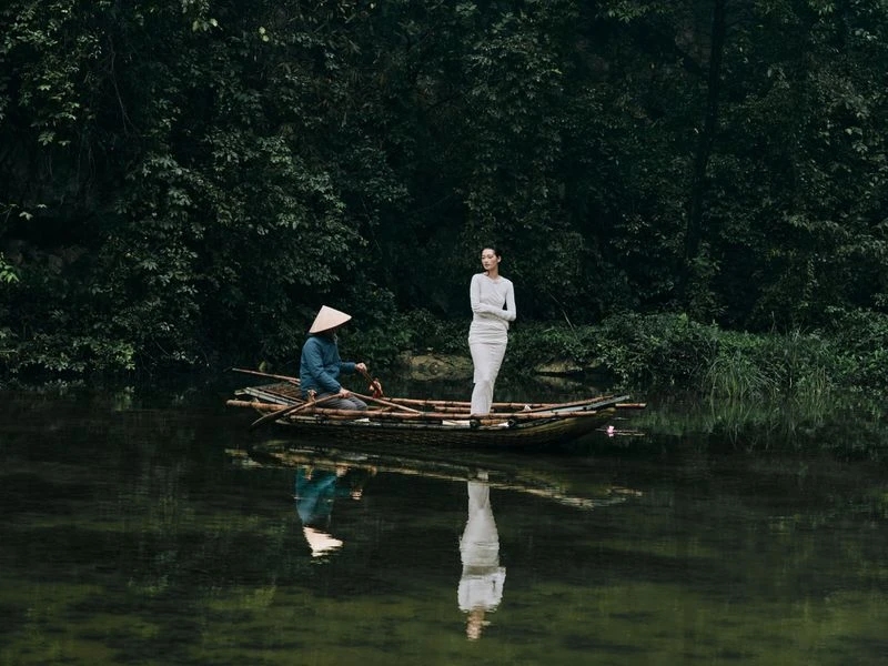Woman in white dress stands in bamboo boat on water near seated man wearing a conical hat in a lush, green, outdoor setting.