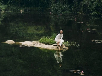 Model in white clothing crouched on a grassy rock outcrop in the middle of a dark river surrounded by lush forest.