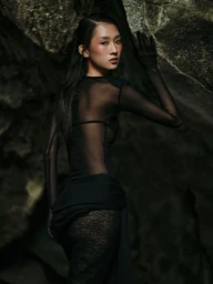 Model in sheer black long-sleeve top and gloves posing against a dark rocky cave wall