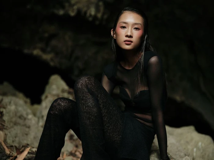 Model wearing a black sheer mesh top and textured pants seated on rocky terrain in a cave environment.