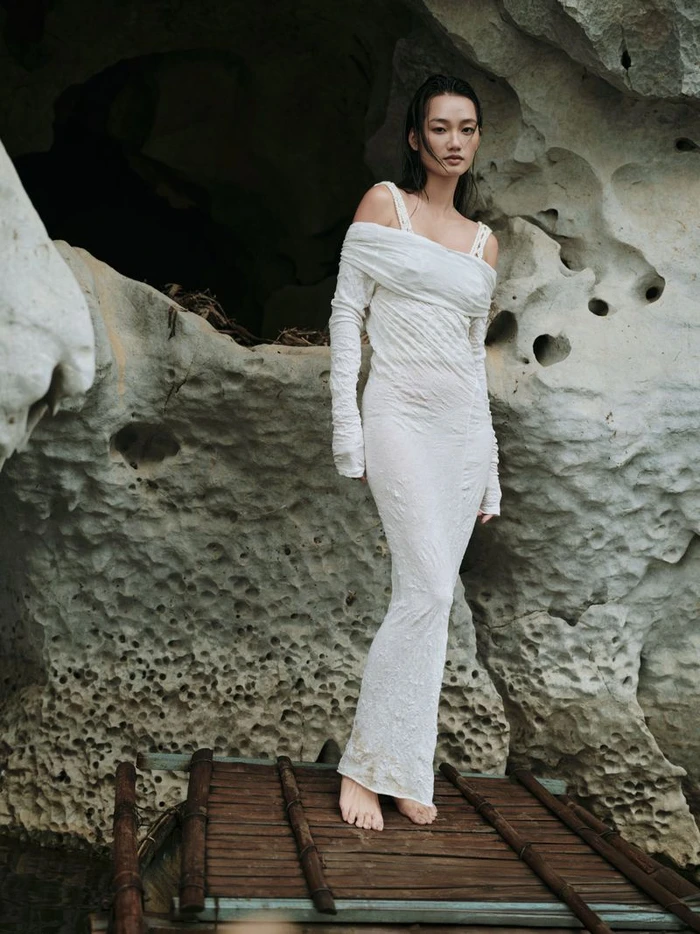 Model wearing a textured white off-the-shoulder dress standing on a wooden dock against a rocky cave background.