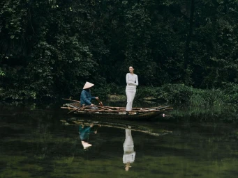 An Asian woman in a long white dress standing on a wooden boat steered by a person wearing a conical hat on a calm river.