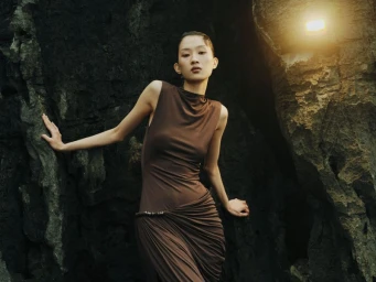 Model in a brown sleeveless dress posing against a dark rocky cave wall with a warm overhead light source.