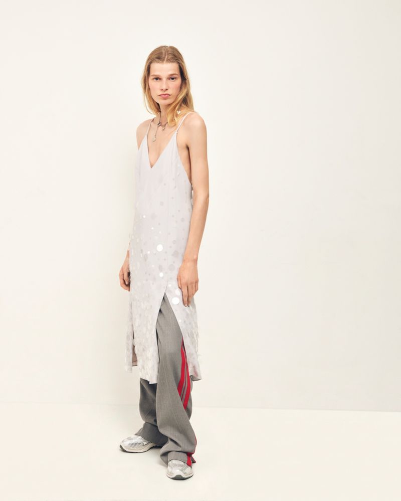 Model wearing a pale embellished slip dress over wide-leg grey track pants with red stripes against a plain studio background.