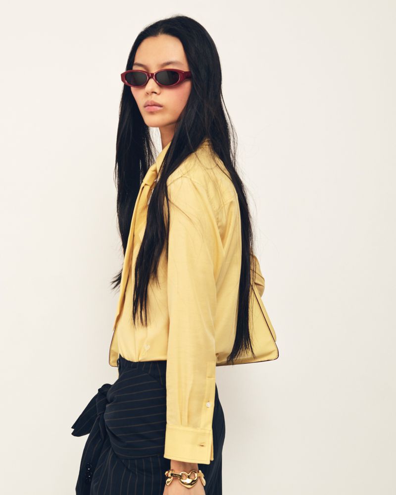 Model with long black hair wearing dark sunglasses and a pale yellow buttoned shirt against a white backdrop.