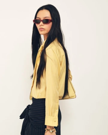 Model with long black hair wearing dark sunglasses and a pale yellow buttoned shirt against a white backdrop.