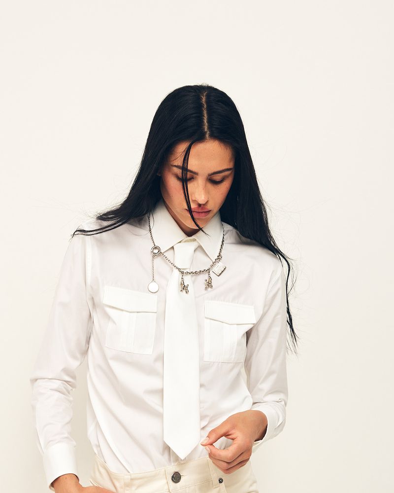 Amelia Gray wearing a white button-up shirt, matching white tie, and silver charm necklace against a plain white studio backdrop.