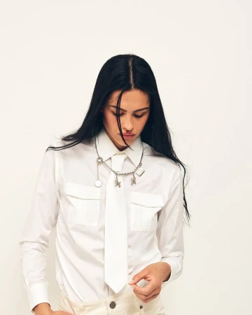Amelia Gray wearing a white button-up shirt, matching white tie, and silver charm necklace against a plain white studio backdrop.