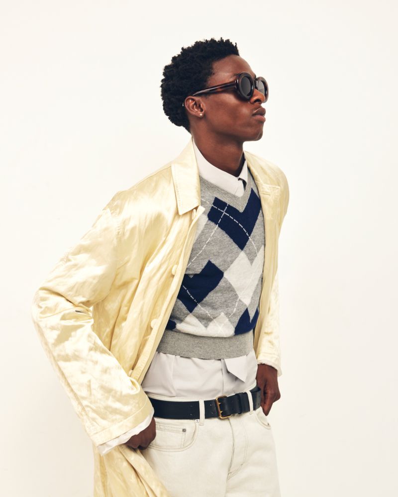 Black male model wearing dark round sunglasses, a pale yellow satin coat, and an argyle knit vest against a white studio backdrop.