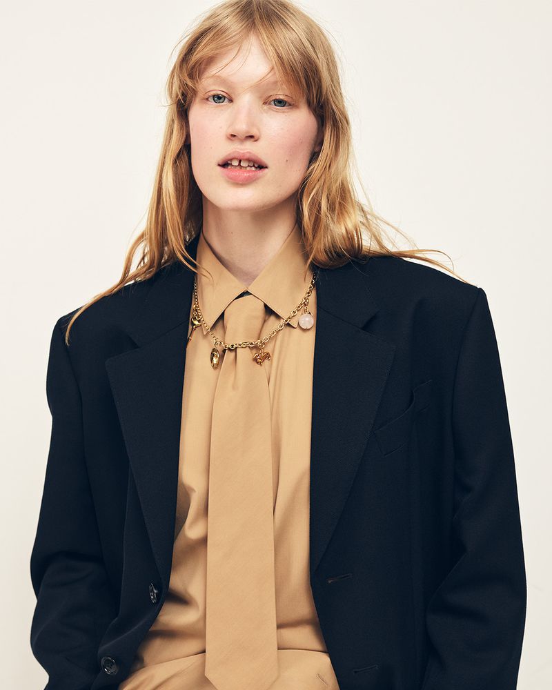 Model wearing an oversized black blazer with a tan shirt, matching tie, and gold charm necklace against a white background.