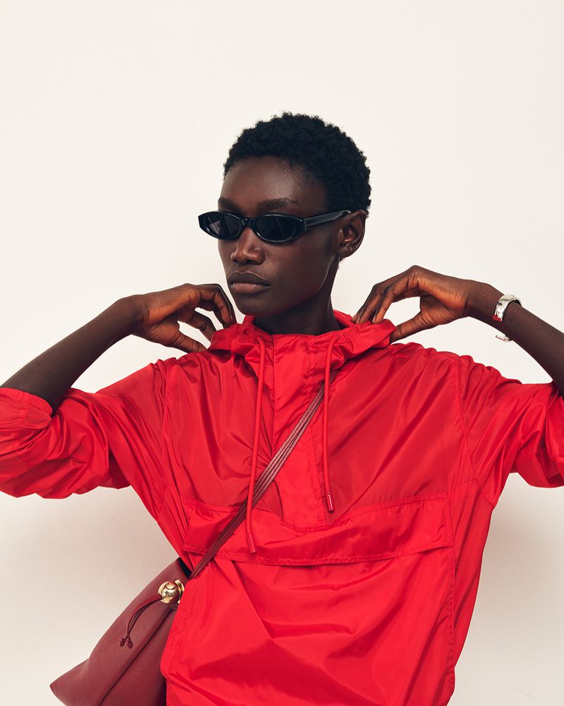 Black model in dark sunglasses wearing a vibrant red hooded windbreaker against a white studio background.