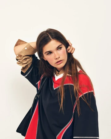 Model posing with one hand behind her head in an oversized navy and red colorblock top over a tan shirt against a white backdrop.