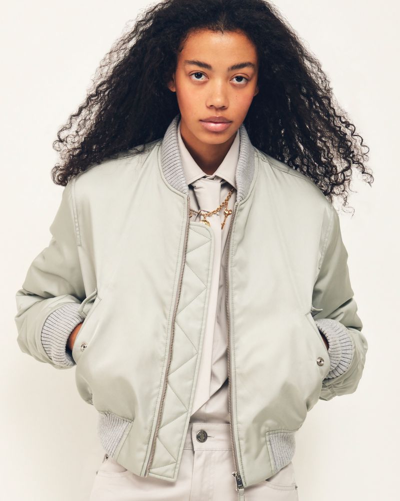 Model with voluminous curly dark hair wearing a pale satin bomber jacket over a collared shirt and tie against a white background.