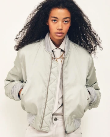 Model with voluminous curly dark hair wearing a pale satin bomber jacket over a collared shirt and tie against a white background.