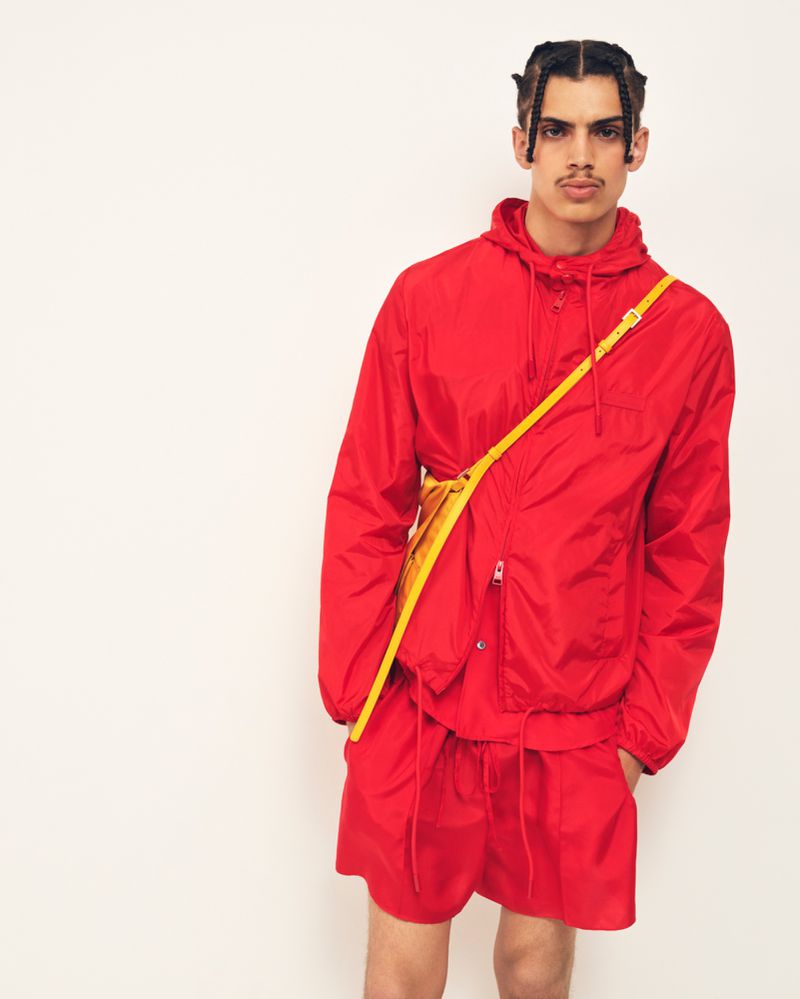 Model wearing a red hooded jacket and red shorts with a yellow strap bag against a white background