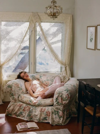 Valerie Pasker reclines in a floral armchair wearing pale pink lingerie and stockings beside a sunlit curtained window.