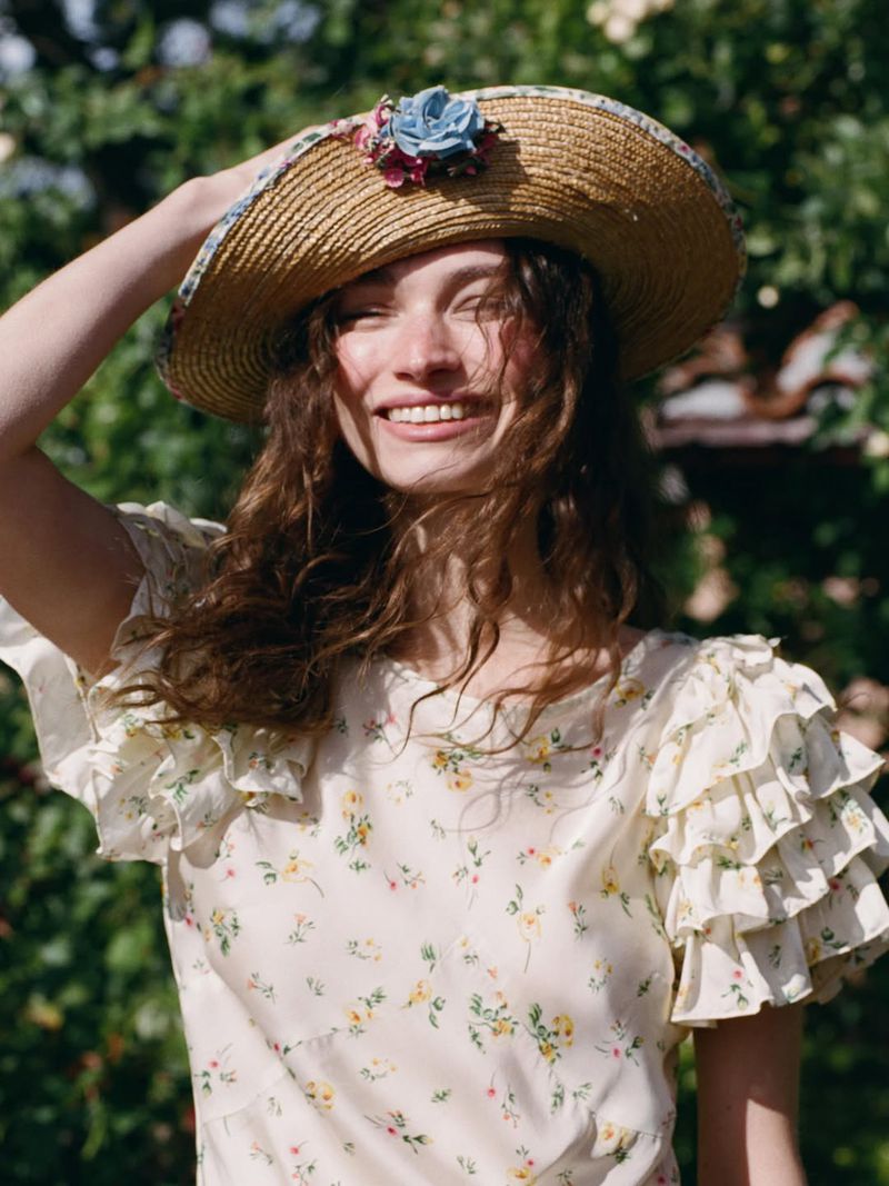 Model Valerie Pasker smiling in a floral ruffle dress and straw hat embellished with a blue flower outdoors in natural sunlight.