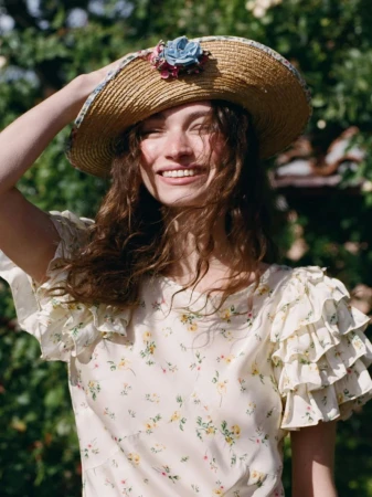 Model Valerie Pasker smiling in a floral ruffle dress and straw hat embellished with a blue flower outdoors in natural sunlight.