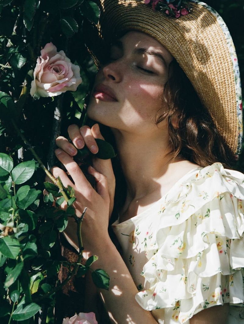 Valerie Pasker wearing a straw hat and ruffled floral dress leans against a rose bush in dappled sunlight.