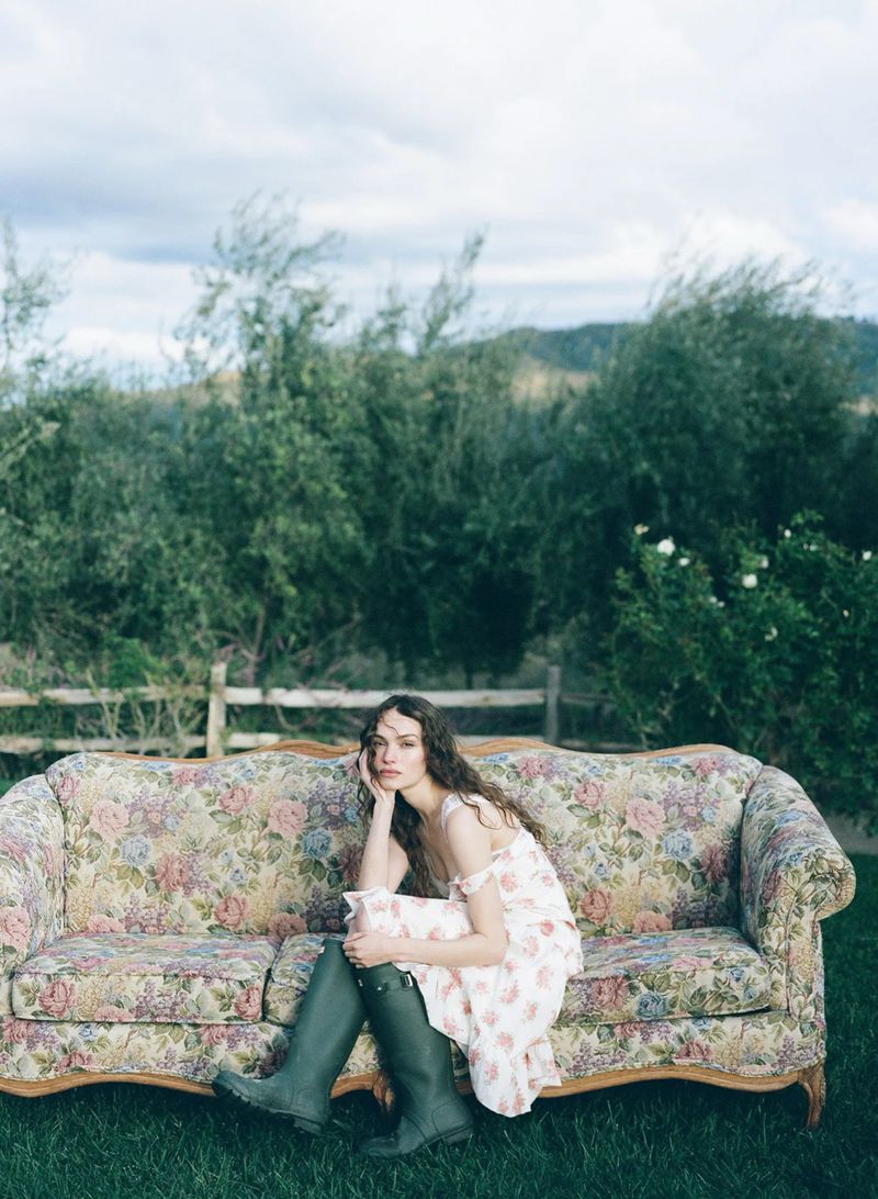 Model Valerie Pasker sitting on a vintage floral sofa in a field wearing a floral dress and dark green rain boots.