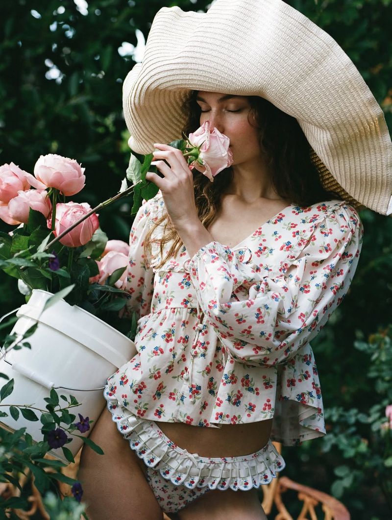 Model wearing a large straw hat smelling a pink rose while holding a bucket of flowers