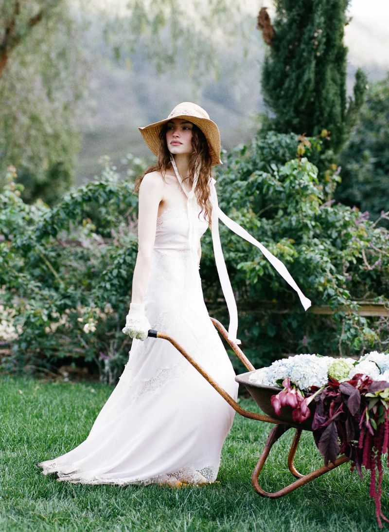 Model Valerie Pasker in a white lace dress and straw hat pushing a wheelbarrow filled with flowers across a lawn.