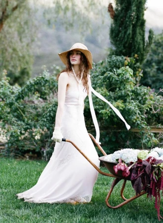 Model Valerie Pasker in a white lace dress and straw hat pushing a wheelbarrow filled with flowers across a lawn.