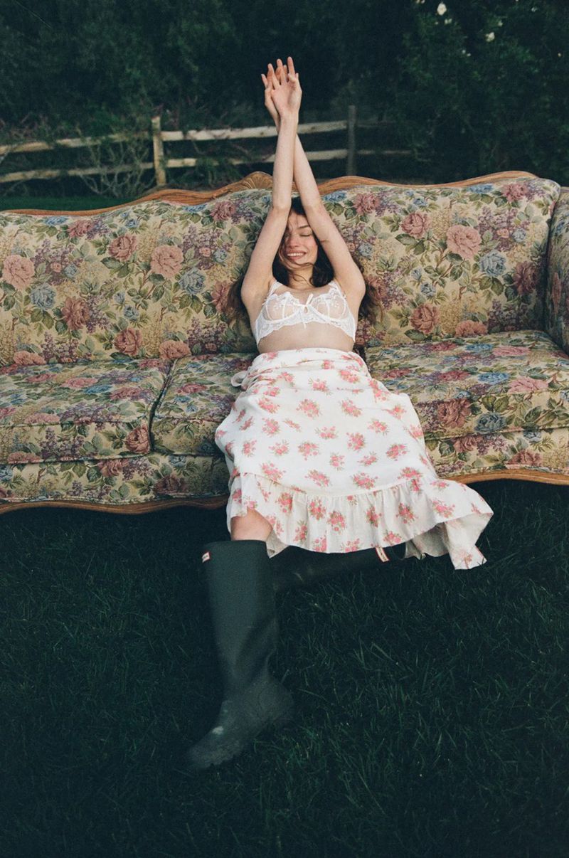 Model Valerie Pasker stretches on a vintage floral sofa outdoors wearing a white lace bralette, floral skirt, and rubber boots.