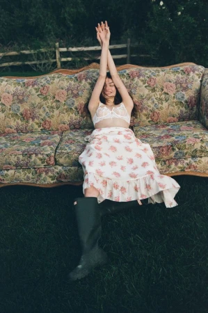 Model Valerie Pasker stretches on a vintage floral sofa outdoors wearing a white lace bralette, floral skirt, and rubber boots.