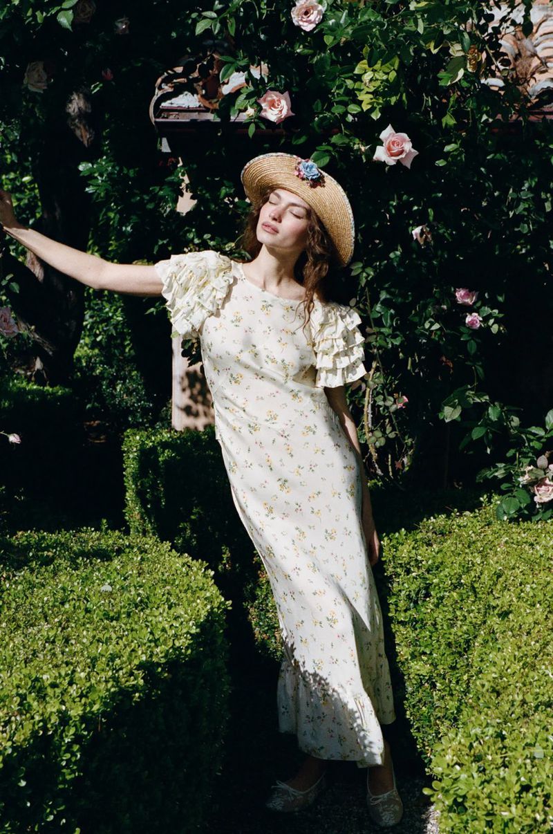 Model Valerie Pasker in a cream floral maxi dress and straw hat posing in a lush rose garden.