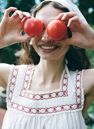 Woman smiling and holding two red tomatoes over her eyes while wearing a white embroidered dress and matching headscarf.