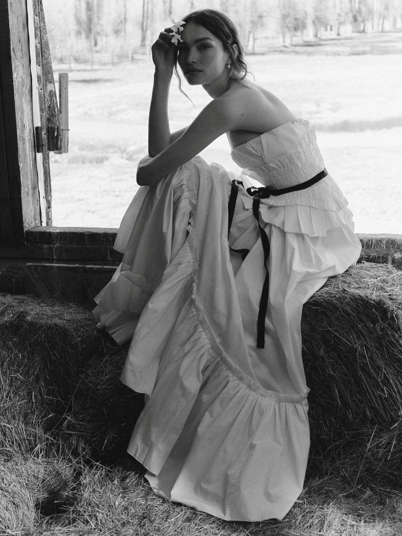 Valerie Pasker wearing a strapless tiered white dress, seated on hay bales beside an open barn window in black and white.