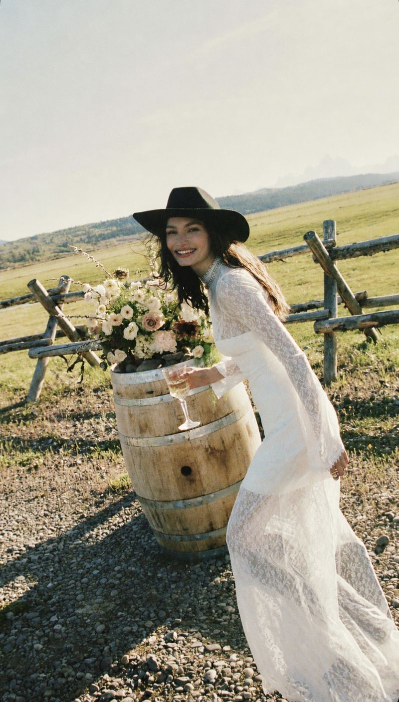 Valerie Pasker wearing a white lace wedding dress and black wide-brim hat holding a wine glass next to a wooden barrel.