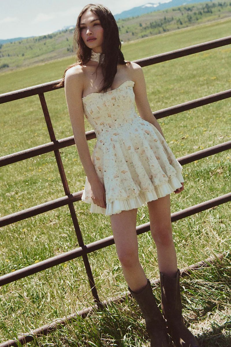 Model Valerie Pasker wearing a strapless floral tiered mini dress and brown boots standing by a metal ranch fence in a green pasture.