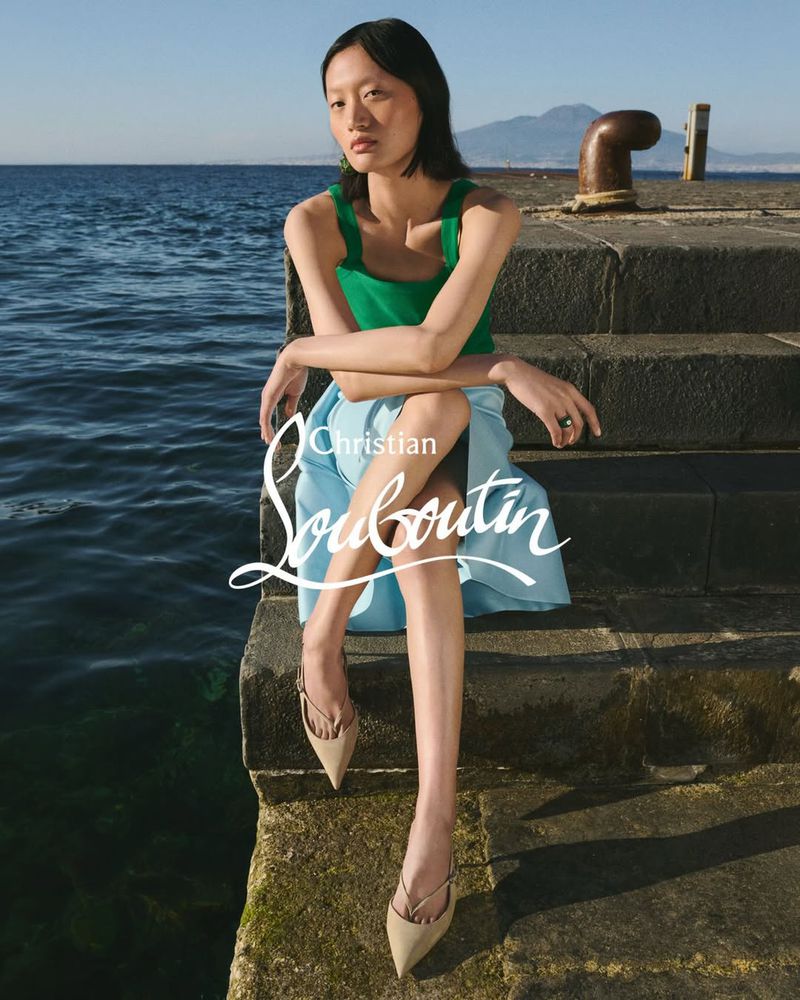 Model seated on stone steps by the ocean wearing a green top and light blue skirt with pointed slingback heels.