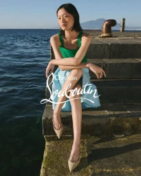 Model seated on stone steps by the ocean wearing a green top and light blue skirt with pointed slingback heels.