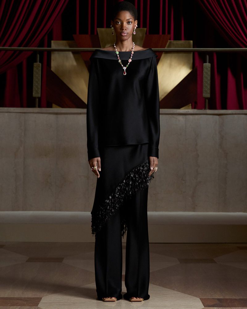 Model in a black off-the-shoulder satin ensemble wearing a Dior high jewelry gemstone necklace against red velvet curtains.