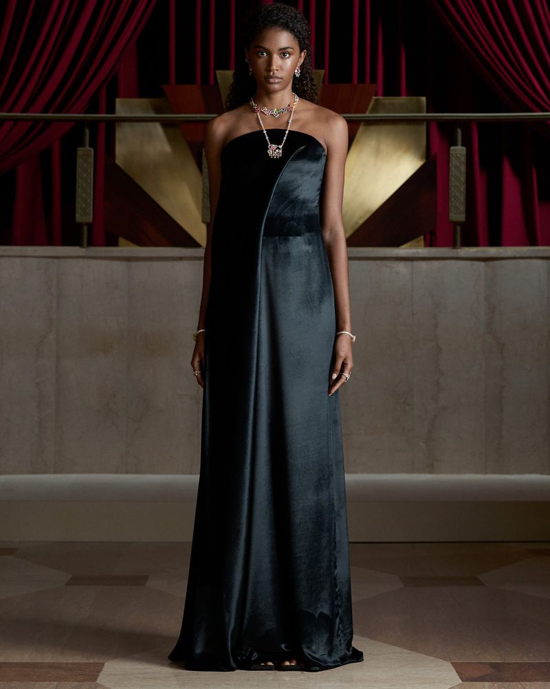 Model wearing a strapless dark satin evening gown and ornate high jewelry necklace standing in front of red velvet curtains.