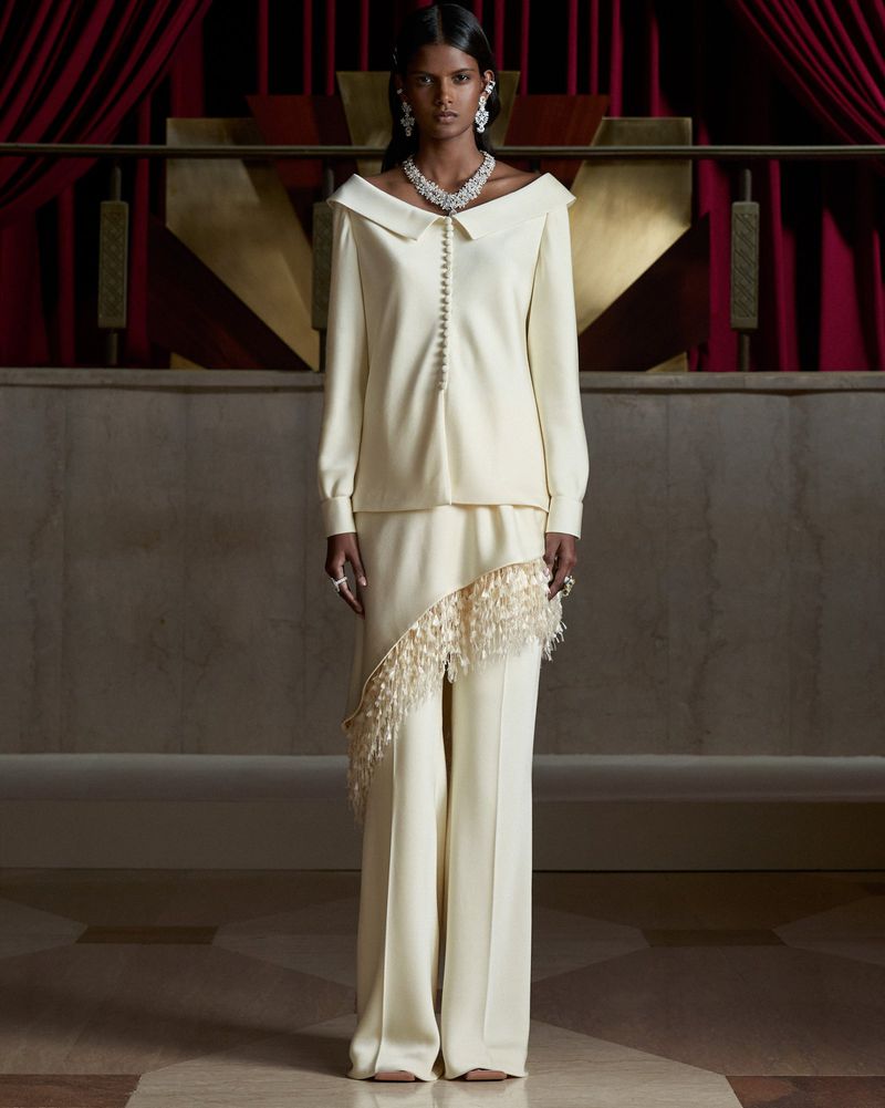 Model wearing an off-white collared suit with fringe-trimmed asymmetrical overskirt and diamond jewelry against red drapery.