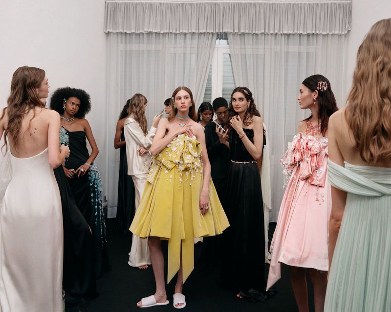 Models backstage wearing couture gowns and high jewelry, centered on a model in an ornate yellow bow dress with slide slippers.